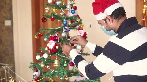 Man wearing Santa hat decorating christmas tree at home during pandemic, Stock Footage 144425411