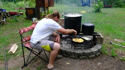 Man wearing shorts sitting next to fire pit watching and checking his food Video stock 249135523