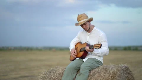 Man wearing a straw hat playing the guitar about a haystack Stockbeeldmateriaal 159899176