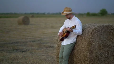 Man wearing a straw hat playing the guitar about a haystack Stock Footage 159899759