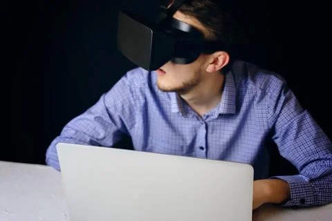 A man is wearing a virtual reality headset while working on a laptop at his desk Stock Photos