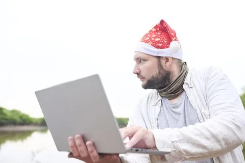 Man wears a Christmas hat while using a laptop outdoors, in nature. Stock Photos