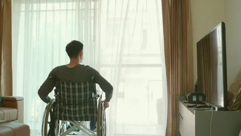 A man in a wheelchair is sitting in front of a window, looking at his cell phone Stock Footage 314449992