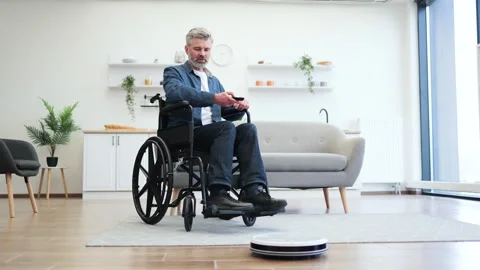 Man in wheelchair using robot vacuum cleaner for efficient home cleaning Stock Footage 317287917