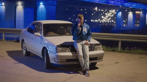 Man by white car under glowing bridge, Contemplative figure leaning on sedan Stock Footage 327352858
