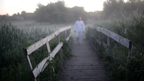 A man in white clothes in out of focus walks along a wooden bridge over a river Stock Footage 221022336
