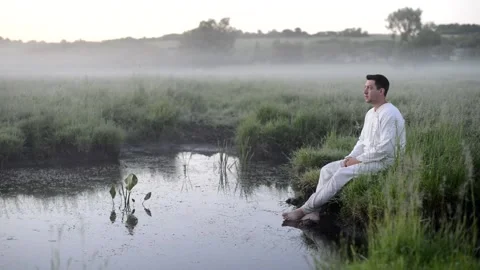 A man in white clothes sits on the grass on the shore of a small forest lake Stock Footage 221022328