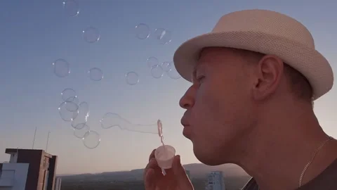 A man in a white hat blowing soap bubbles, against the sky at sunset Stock Footage 282119816