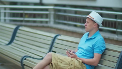 A man in a white hat is resting while sitting on a city bench, tracking a camera Stock Footage 148113679
