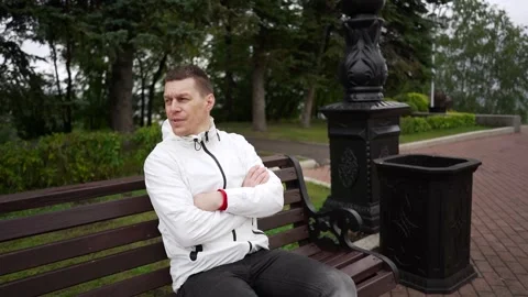 A man in a white jacket is sitting on a bench in the park and watching if it is Stock Footage 266442127