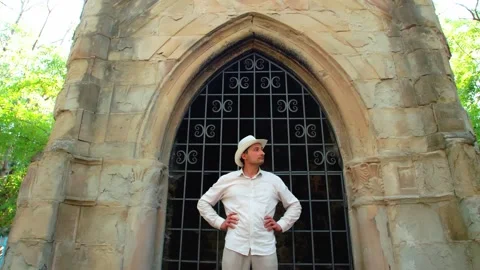 A man in a white straw hat stands in front of an old Gothic building and turns Stock Footage 245483018