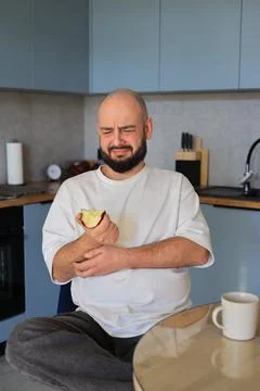 Man wincing while holding a bitten apple in a kitchen Foto stock