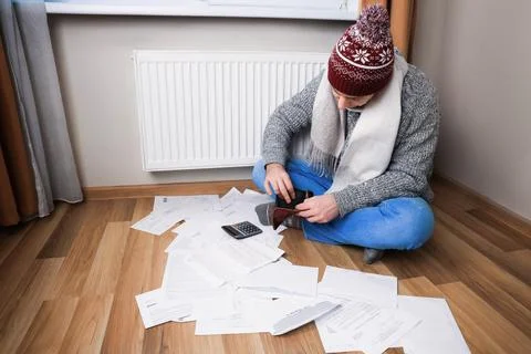 Man in winter clothes checking empty wallet near radiator surrounded by bil.. Stock Photos