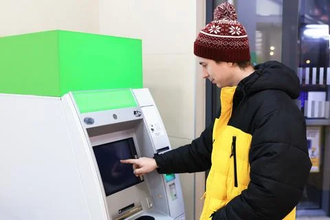 Man in winter clothes using ATM with green mockup screen. Person withdrawin.. Stock Photos