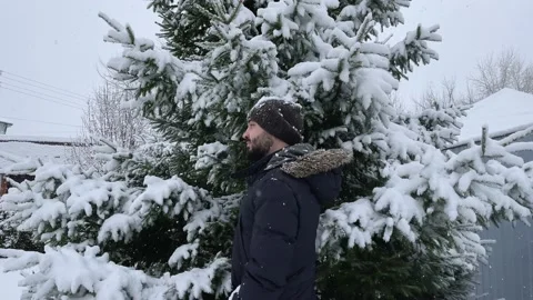 Man in a winter jacket stands under the branches of a Christmas tree. Snow falls Stock Footage 221713920