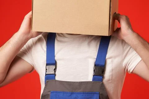 Man in work clothes on a red background put a cardboard box on his head Stock Photos