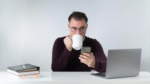 A man at work drinks coffee and looks at the phone screen in surprise with big Stock Photos