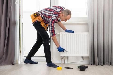 Man in work overalls using wrench while installing heating radiator in room Stock Photos