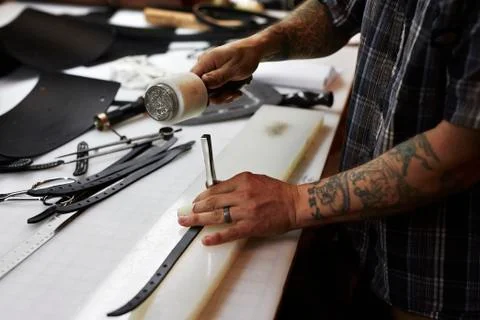 A man working at a bench in a leather workshop, using a mallet and chisel to Stock Photos