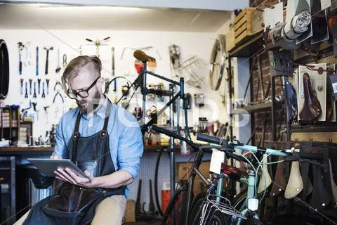 A man working in a bicycle repair shop, seated using a digital tablet. Stock Photos