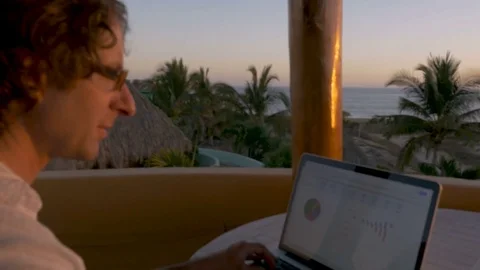 Man working on computer looking at charts during sunrise or sunset at beach Stock Footage 80137000