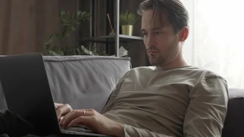 A man is working from home, lying on the couch and typing on his laptop Stock Footage 253403263