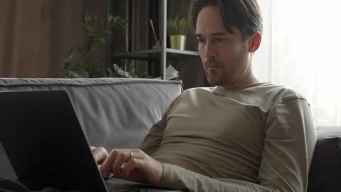 A man is working from home lying on the couch, thinking and typing on his laptop 스톡 동영상 253403312