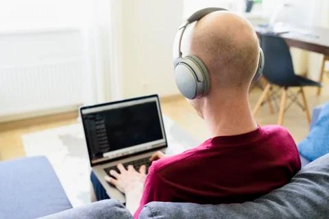 Man working from home on sofa. Using laptop and headphones. Coding, programmi Stock Photos