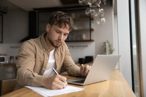 Man working from home writing up data from laptop screen Stock Photos