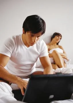 Man working on laptop computer in bed, woman in background. Stock Photos