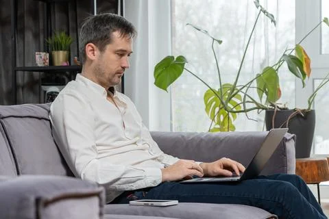 Man working on laptop computer while sitting on gray sofa in living room 스톡 사진