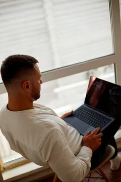A man is working on a laptop by a large window. Man sitting in chair with lap Stock Photos