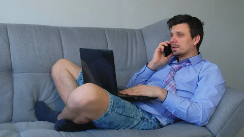 Man working on laptop talking smartphone at home sitting on couch distant work. Stock Footage 128963686