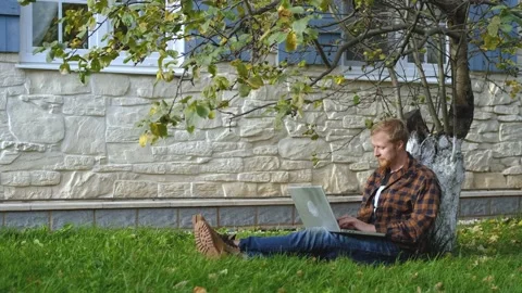 Man working on a laptop while sitting under a tree near the house Stock Footage 200826126