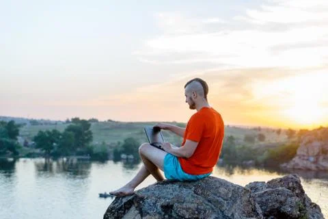 A man working on a laptop while sitting on a rock on a beautiful sunset. Stock Photos