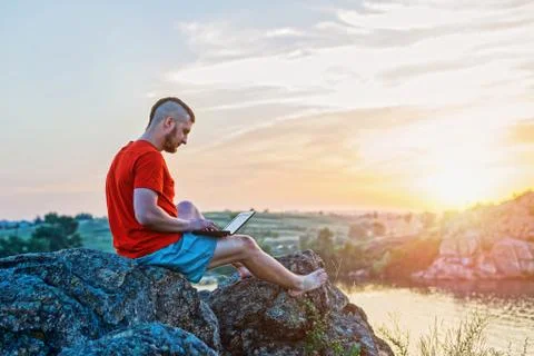 A man working on a laptop while sitting on a rock on a beautiful sunset. Stock Photos