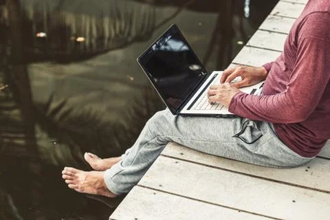 Man working on laptop while sitting on wooden dock. legs in river water Stock Photos