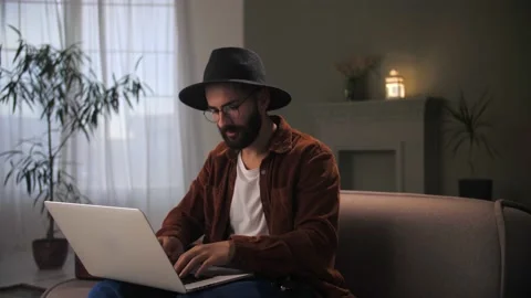 Man working remotely using laptop sitting at couch.Young guy with hat eyeglasses Stock Footage 212316627