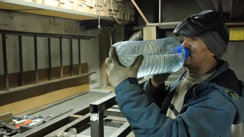 A man working in a workshop drinks water from a large plastic bottle. Hard Stock Footage 322733670