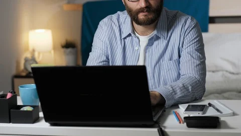 Man works on computer and drinks coffee. Bearded guy sits at desk in bedroom Stock Footage 129376296