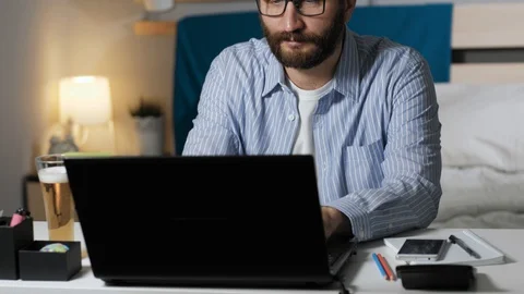 Man works on computer and drinks beer. Bearded guy sits at desk in bedroom Stock Footage 129377078