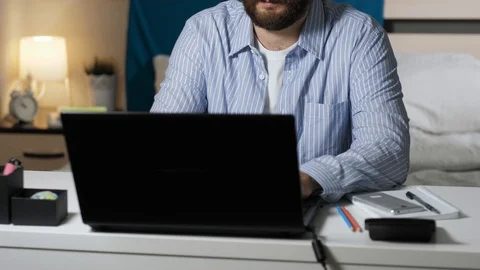 Man works at computer at home. Bearded guy sits at table in bedroom and types on Stock Footage 129371929