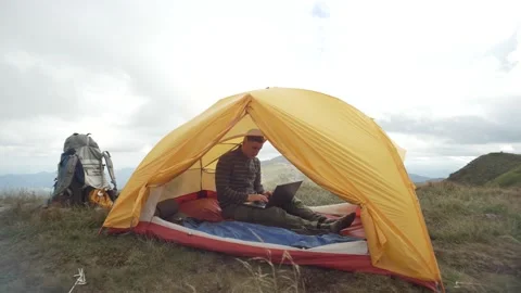 A man works at a computer while sitting in a tent with a mountain landscape. Stock Footage 213017222
