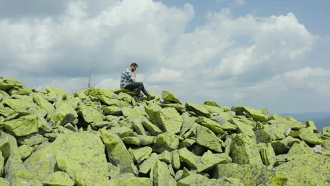 A man works on a laptop while sitting on top of a rocky mountain. Stock Footage 248203023