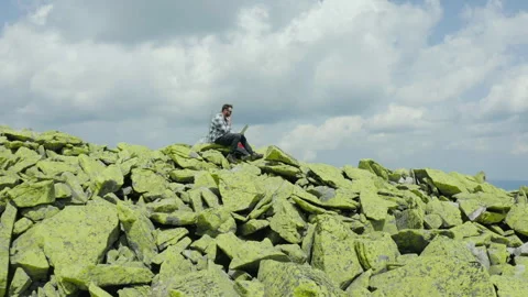 A man works on a laptop while sitting on top of a rocky mountain. Stock Footage 248203285