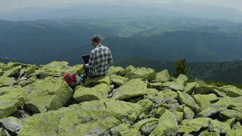A man works on a laptop while sitting on top of a rocky mountain. Stock Footage 248203708