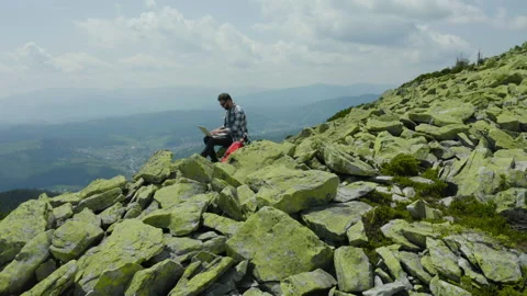 A man works on a laptop while sitting on top of a rocky mountain. Stock Footage 248203780