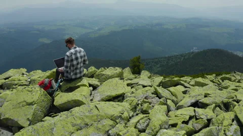 A man works on a laptop while sitting on top of a rocky mountain. Stock Footage 248203805
