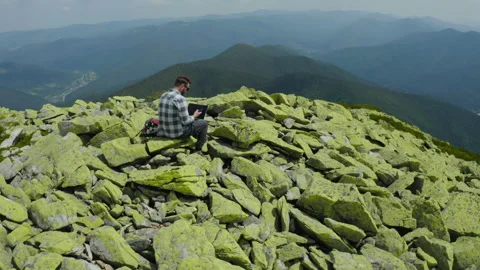 A man works on a laptop while sitting on top of a rocky mountain. Stock Footage 248204124