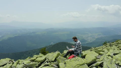 A man works on a laptop while sitting on top of a rocky mountain. Stock Footage 248204659